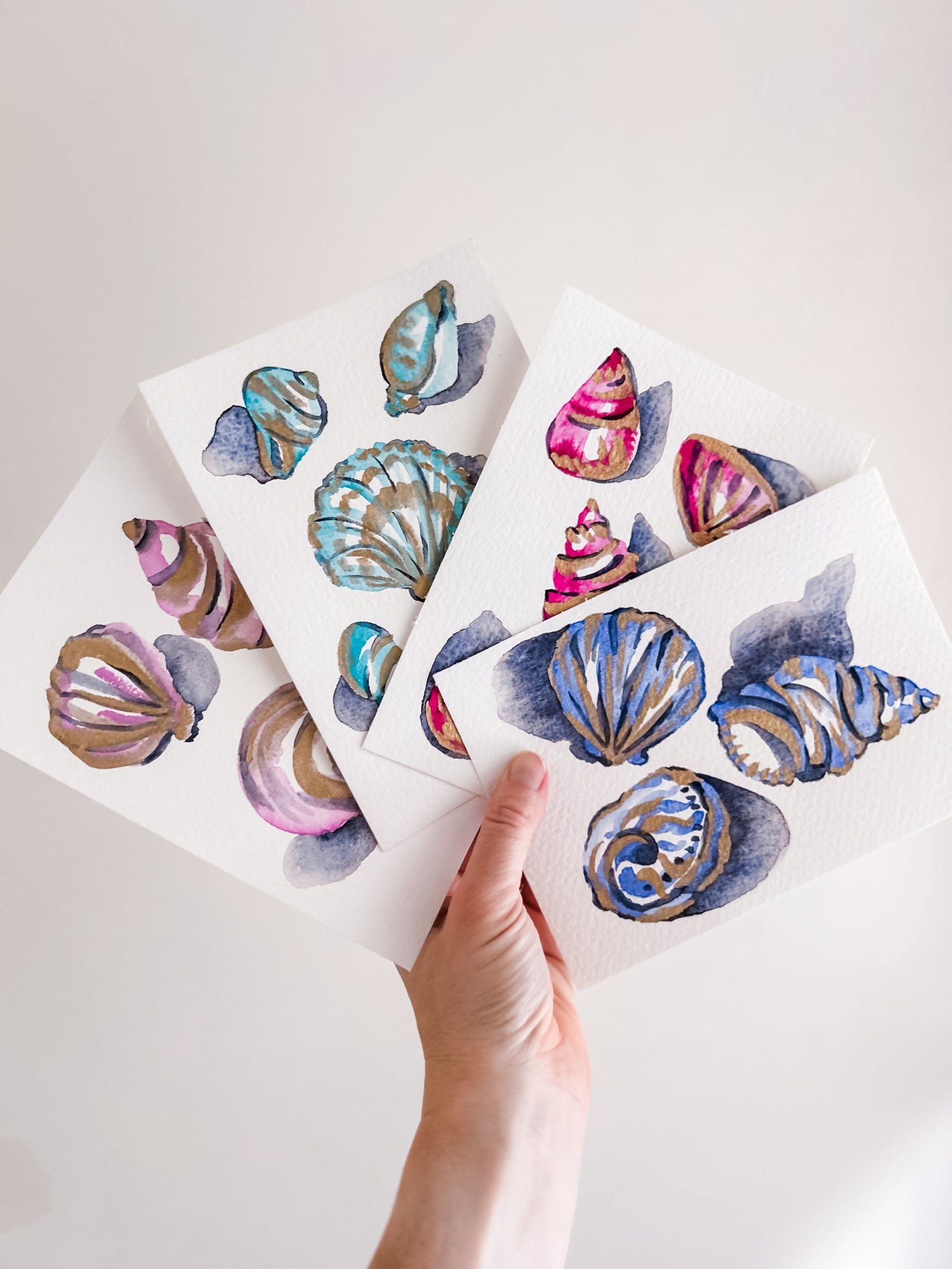 A hand holding up four watercolor paintings of seashells in pink, teal, and blue